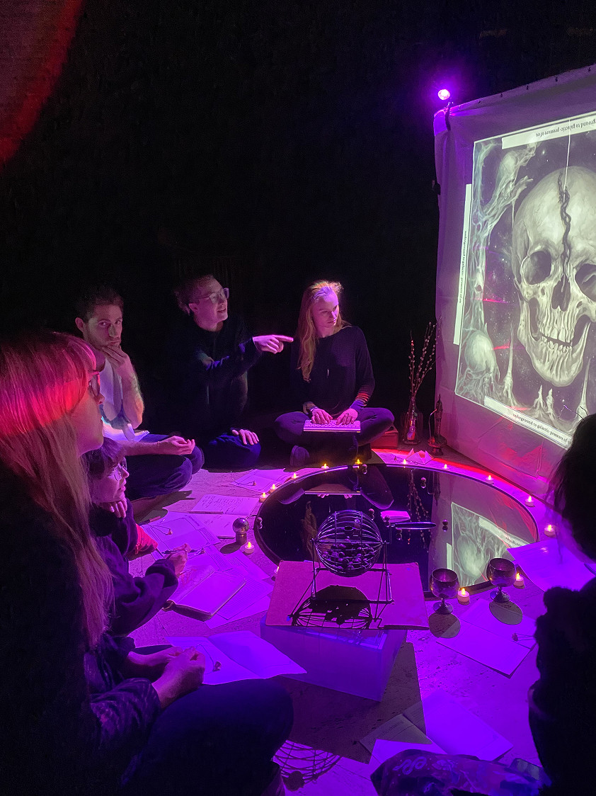 A group sits around a lit-up mirror and discusses a projected image of a skull